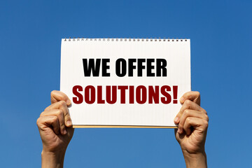 We offer solutions text on notebook paper held by 2 hands with isolated blue sky background. This message can be used as business concept about offering solutions.