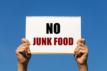 No junk food text on notebook paper held by 2 hands with isolated blue sky background. This message can be used as business concept to encourage audience not to eat junk food.