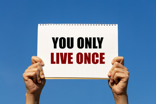 You only live once text on notebook paper held by 2 hands with isolated blue sky background. This message can be used as business concept to encourage audience to live your life.