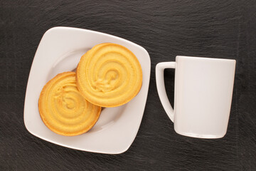 Two sweet cookies on white ceramic saucer with cup on slate stone, macro, top view.