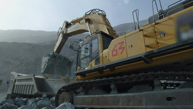 Yellow opencast excavator loading ore into the body of heavy tipper, which driving in reverse in dusty quarry. Excavator and a dump truck working in an open pit