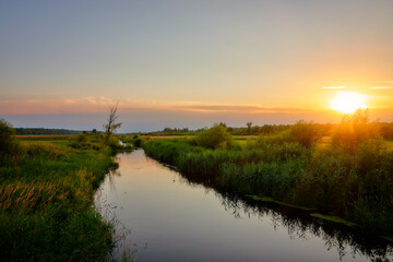 Fototapeta na wymiar River runs through a grassy field at sunset.