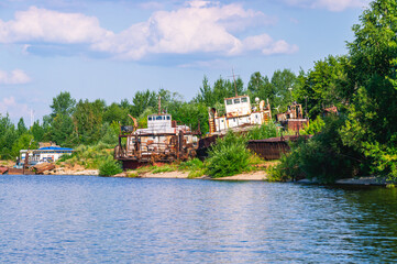 Old rusty ships lie on the riverbank. The river bank and rusting ships. Pier with old ships on the shore. Old tugs are rusting on the riverbank.