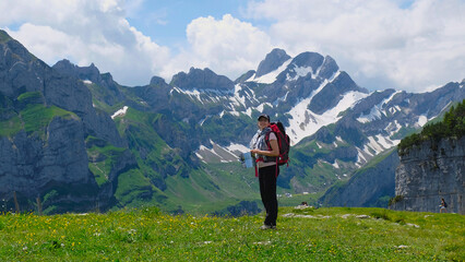 Woman with hat and backpack holds a touristic leaflet and smiles while being surrounded by an alpine landscape, snowy peaks and green pastures. Appenzell, Switzerland