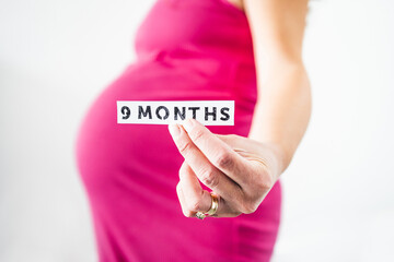 pregnant woman holding 9 Months sign towards the camera wearing pink dress in the last month of pregnancy