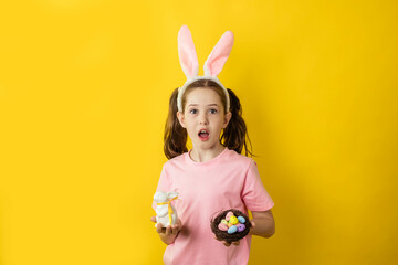 Cute surprised teenage girl on a yellow background in a rabbit costume holds in her hands an Easter...