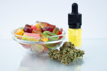 Assorted different gummy candies in a small glass bowl and a bottle of CBD canna oil, near in the foreground several buds of dried medical marijuana, reflected on the glass