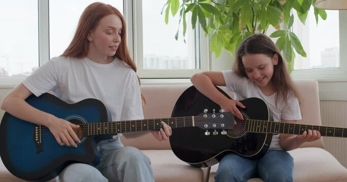 red-haired woman is explaining to student how to play guitar in the bedroom,little adorable girl improves skills with tutors at home slow motion, kid learning Finger, thumb Positions