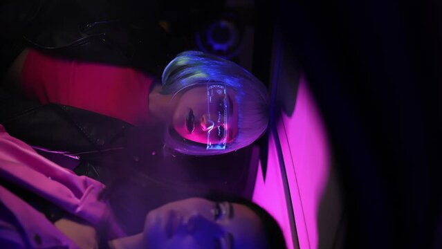 Portrait beautiful trendy woman wear white outfit and futuristic glasses posing looking into camera standing by illuminated wall in night club. Cyberpunk style, sensual look clubber in neon blue light