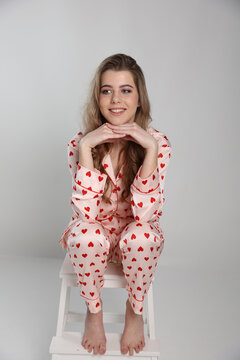 beautiful blonde girl sits on a chair on a white background in pajamas. pajamas with hearts. Valentine's Day. studio photo shoot in satin pajamas with a heart print. clothes for sleep and home