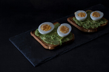 Toast with avocado and boiled egg, on a slate tray, on a dark background. Dark mood.