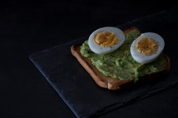 Toast with avocado and boiled egg, on a slate tray, on a dark background. Dark mood.