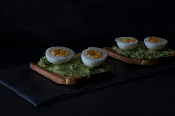 Toast with avocado and boiled egg, on a slate tray, on a dark background. Dark mood.