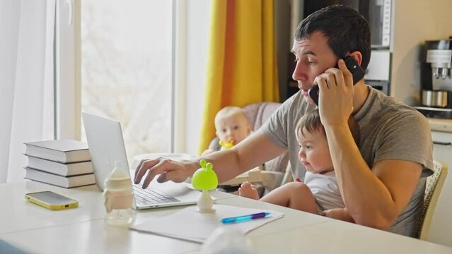 Father Working From Home Remotely With Baby Daughter In His Arms. Pandemic Remote Work Business A Concept. Father Tries To Work At Home In Kitchen, Baby Fun Children Interfere Sitting On Their Hands