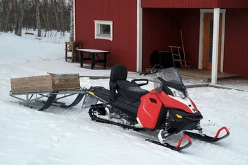 Fotobehang A snowmobile with wooden trailer  stands by wooden Scandinavian red house. © Iwona