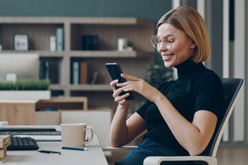 Happy young businesswoman using smart phone while sitting at her working place in office