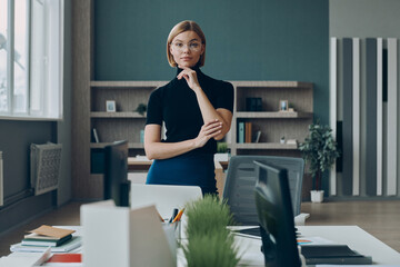 Serious businesswoman looking at camera while standing near her working place in office