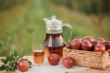 A glass with apple juice and basket with apples on wooden table with natural orchard background. Vegetarian fruit composition