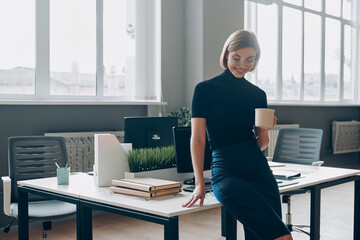 Happy businesswoman holding coffee cup while resting near her working place in office