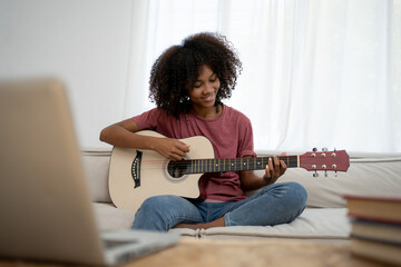 Focused African girl playing acoustic guitar and watching online course on laptop while practicing at home. Online training, online classes. - obrazy, fototapety, plakaty