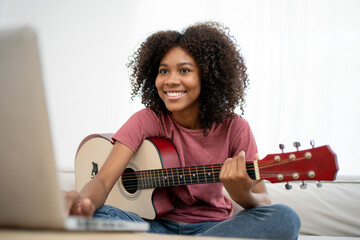 Focused African girl playing acoustic guitar and watching online course on laptop while practicing...