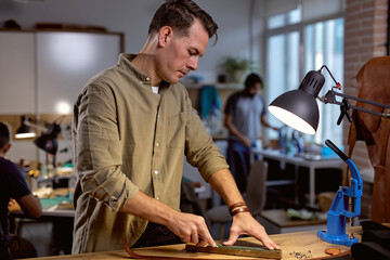 young handsoem craftsman polishes the edge of belt, man sewing leather belt at workplace, side view shot, working people in the background of the photo