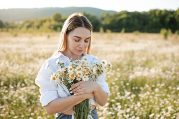 Naklejka na ściany i meble Beautiful attractive blonde woman 25-27 year old pick up chamomile flowers walk in blooming meadow outdoor. Spring season.