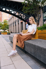 Vertical side view of focused creative hipster female artist drawing in sketchbook enjoying spending free time on hobby. Pensive young woman writing notes and planning recreation on bench outdoors.