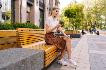 Wide shot of pretty female with long blond hair in casual clothes using smartphone sitting on bench...