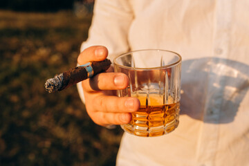 Close-up photo of a man's hand holding a clear glass of whiskey and a Cuban cigarette