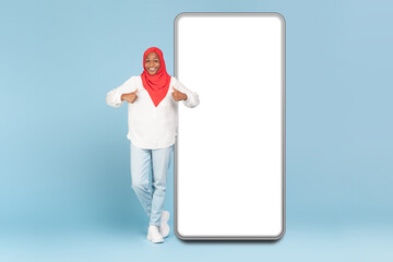 Cheerful black woman in hijab standing near huge new cellphone with white empty screen and showing...