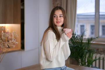 Young beautiful happy woman enjoy with a cup of tea or coffee at kitchen at home in sweater, smile, look at camera