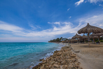 View of Atlantic ocean and beach with sun loungers and umbrellas against backdrop blue sky with white clouds. Aruba.