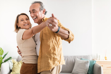 Cheerful retired spouses husband and wife dancing and laughing in living room, happy romantic couple enjoying slow dance