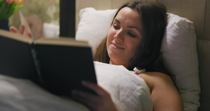 Young woman is reading interesting book while lying in bed
