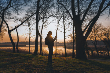 Woman photographer with a camera in her hands and a backpack standing in the spring forest at sunset on the lake