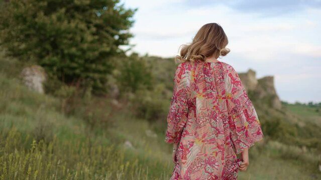 The camera follows a young pretty hipster woman girl in a stylish dress walking on top of a cliff against the backdrop of the river, circling smiling posing for the camera, happy and drunk for life