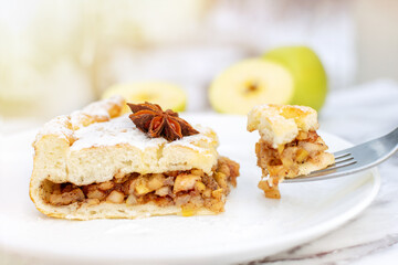A piece of apple pie with cinnamon on plate, breakfast in a cafe on a sunny morning. Breaks off a part of cake sprinkled with powdered sugar and anise star using a fork. Delicious dessert, pastries.
