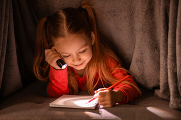 Cute little girl with flashlight hiding under blanket and writing in notebook