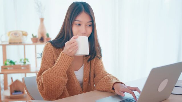 Smiling young asian woman using laptop while holding a cup of coffee at home. lifestyle