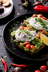 Avocado Toast with Poached Eggs and Cherry Tomatoes on Black Plate.
Healthy breakfast toast topped with smashed avocado, poached eggs, cherry tomatoes and chives, served with lemon on a dark table.