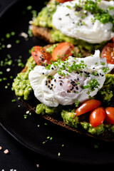 Avocado Toast with Poached Eggs and Cherry Tomatoes on Black Plate.
Healthy breakfast toast topped with smashed avocado, poached eggs, cherry tomatoes and chives, served with lemon on a dark table.