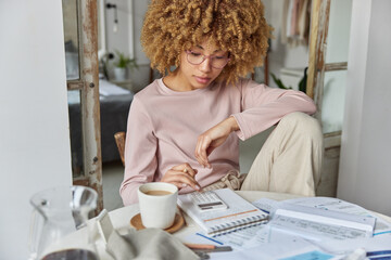 Serious curly haired woman manages household family budget calculates expenditures takes care of finances and savings sits at table with receipts dressed in domestic clothes poses at home office