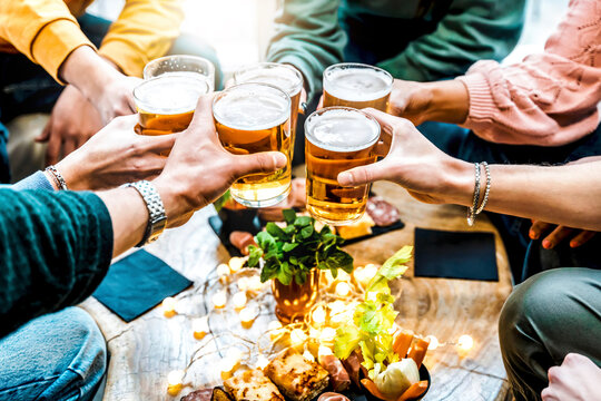 Friends cheering beer glasses on wooden table at brewery bar indoor at patio party - People having fun at happy hour at bar restaurant - Food and beverage lifestyle concept