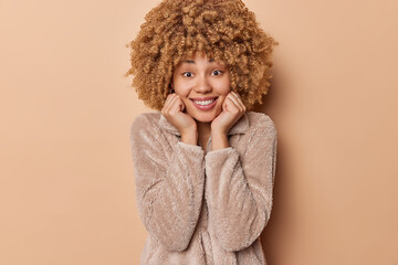 Happy positive woman keeps hands under chin smiles gladfully shows white teeth dressed in winter fur coat expresses sincere emotions isolated over brown background listens something with pleasure