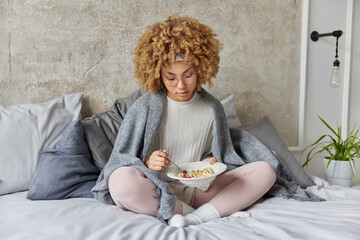 Curly haired woman in pajama has healthy breakfast in morning eats muesli with fruits covered with plaid sits crossed legs on comfortable bed against cozy home interior. People and lifestyle concept
