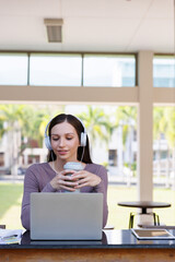 Young female student waering headphones and holding thermos bottle studying with laptop at table in college. Success business woman surfing the net on laptop.