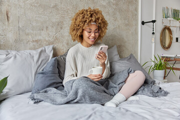 Cheerful relaxed curly woman drinks coffee and surfs internet on smartphone enjoys lazy day at home poses on comfortable bed under blanket against cozy domestic interior enjoys online communication