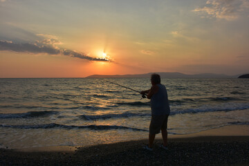 An amateur old fisherman on the beach at a scarlet sunset throws his fishing rod into the sea