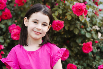 Smiling child girl 5-6 year old with rose flowers over blooming bushes in garden outdoor. Childhood. Spring season.